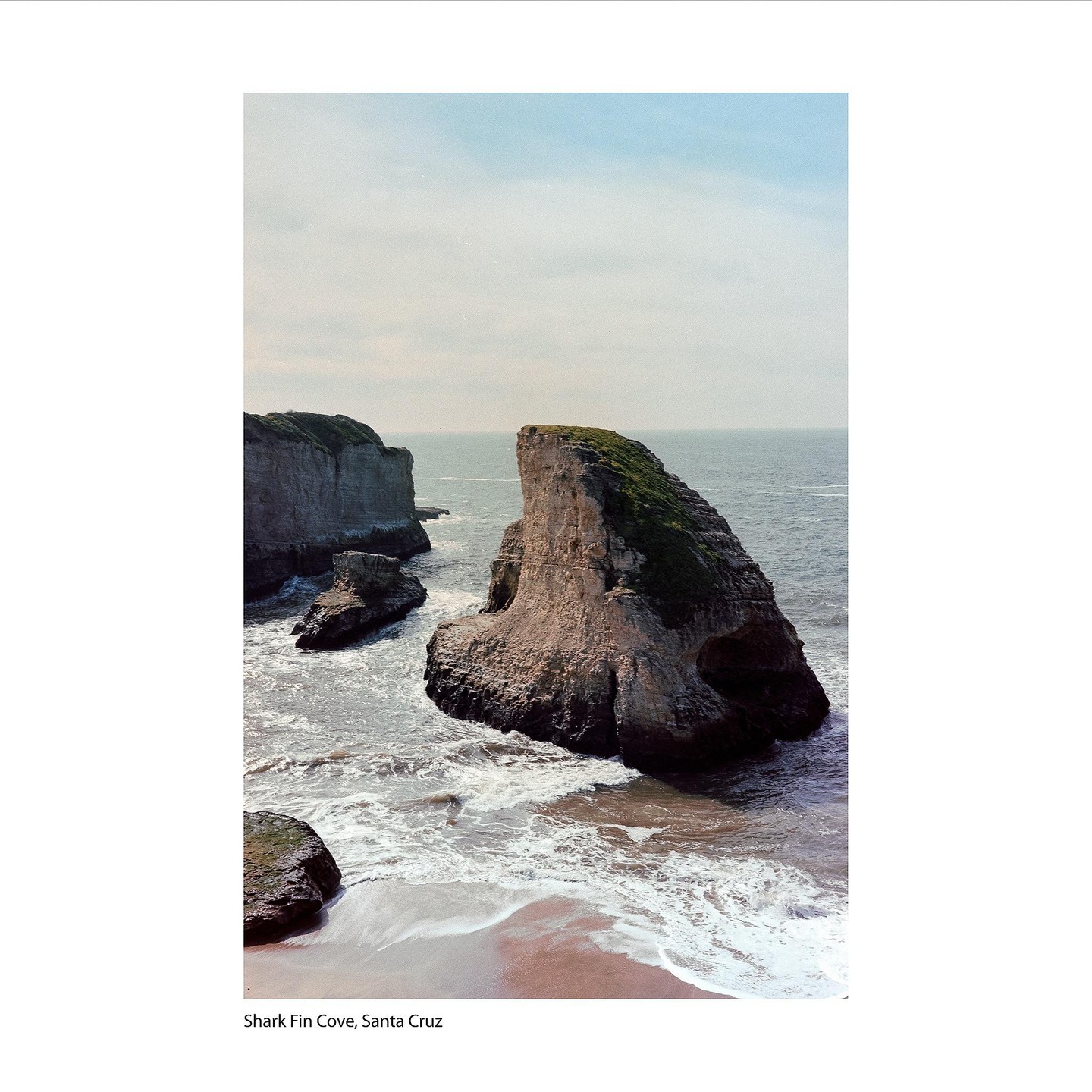 Waves crash against a rocky beach with a large rock formation resembling a shark fin.