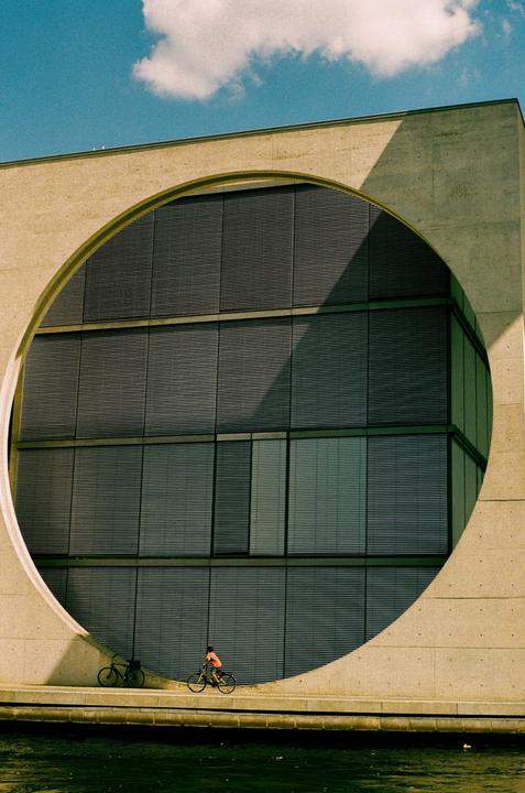 A cyclist rides past a large concrete building with a circular cut-out and many windows.