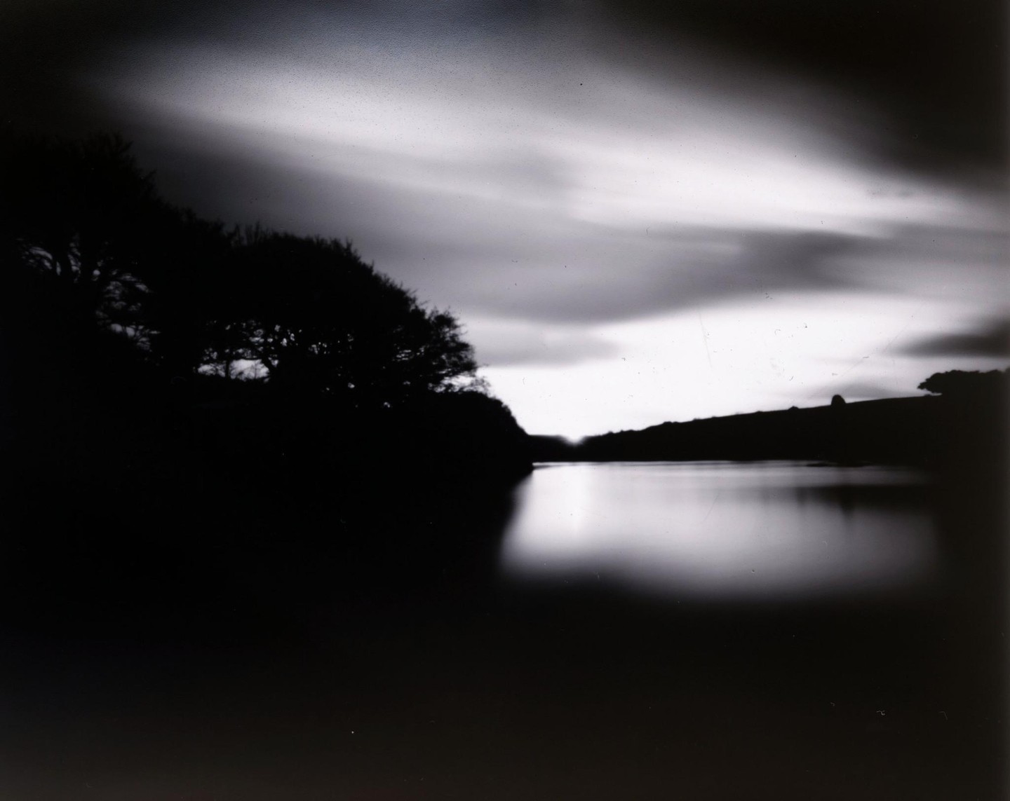 A body of water reflects the sky and a dark tree-lined shore under a cloudy sky.