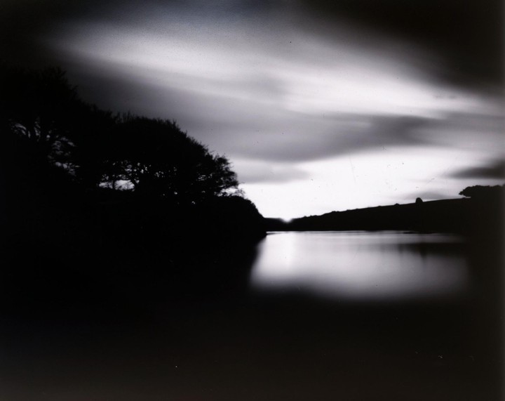 A body of water reflects the sky and a dark tree-lined shore under a cloudy sky.