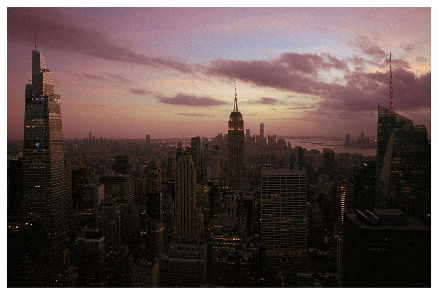A cityscape at dusk shows the Empire State Building illuminated among other skyscrapers under a purple and orange sky.