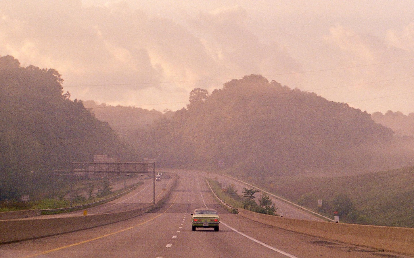 A green car drives away on a highway surrounded by hills under a hazy sky.