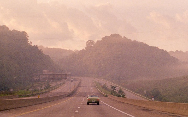 A green car drives away on a highway surrounded by hills under a hazy sky.