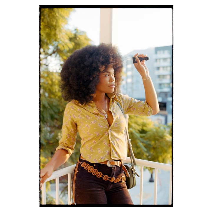 A woman with an afro combs her hair while standing on a balcony.