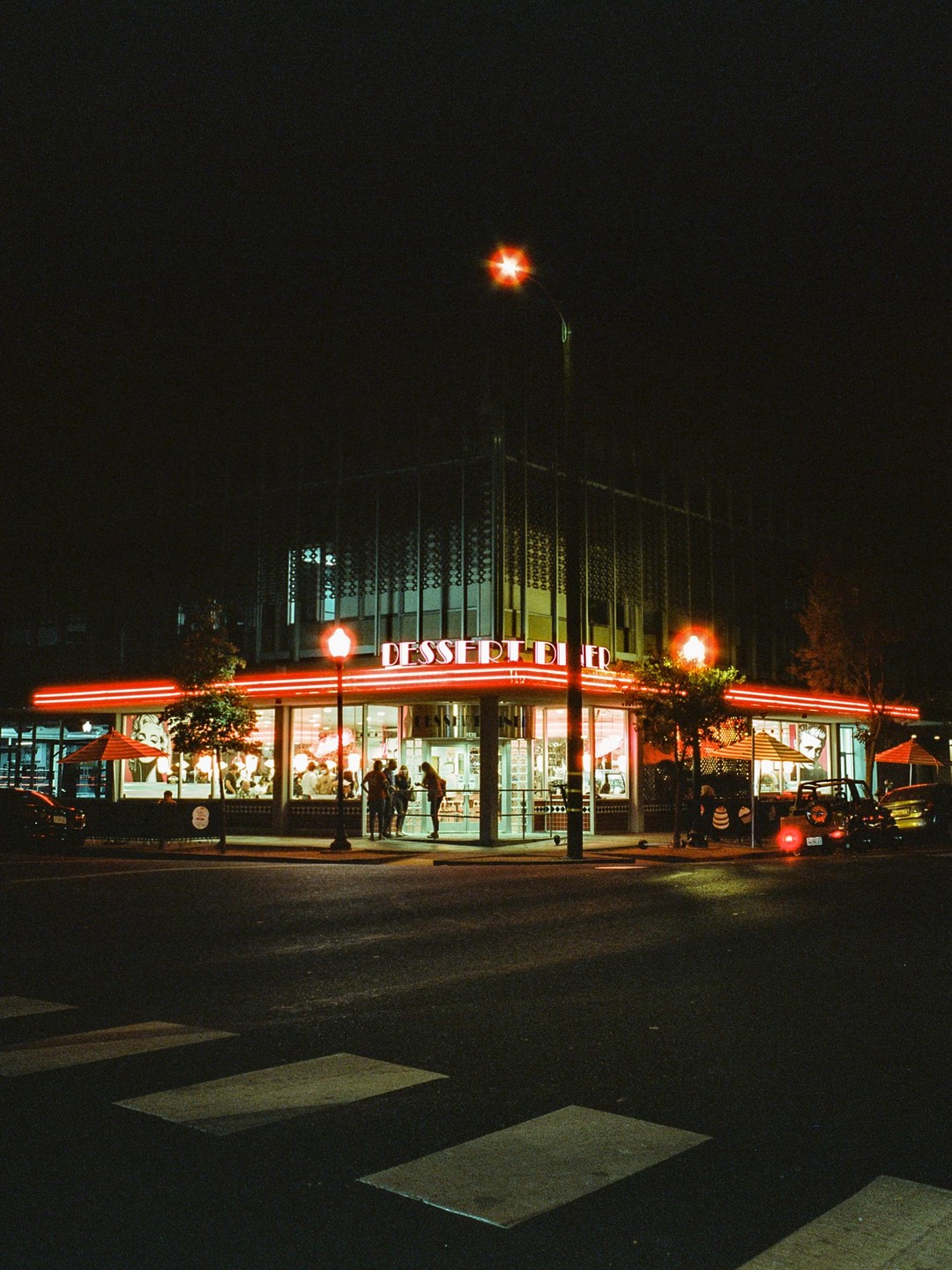 A dessert diner with red neon lights illuminates a street corner at night.