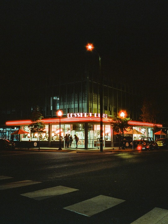 A dessert diner with red neon lights illuminates a street corner at night.