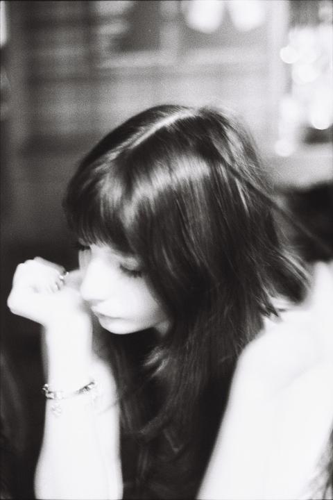 A woman with long hair and bangs looks down, resting her chin on her hand, wearing a bracelet.