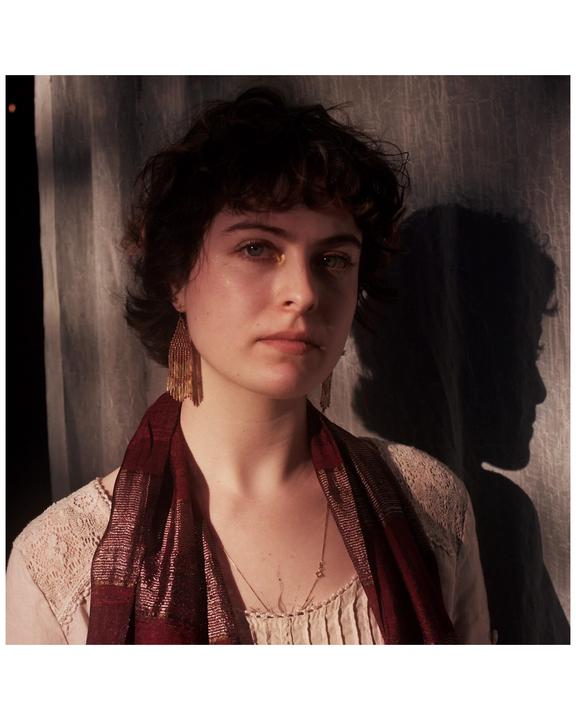 A woman with short, curly hair and a red scarf looks directly at the viewer, with her shadow cast on the wall behind her.