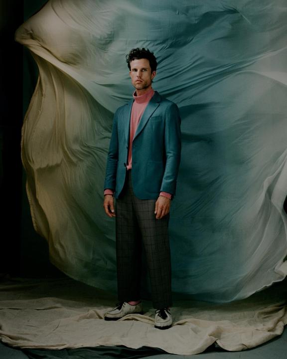 A man in a teal jacket and pink turtleneck stands in front of a flowing fabric backdrop.