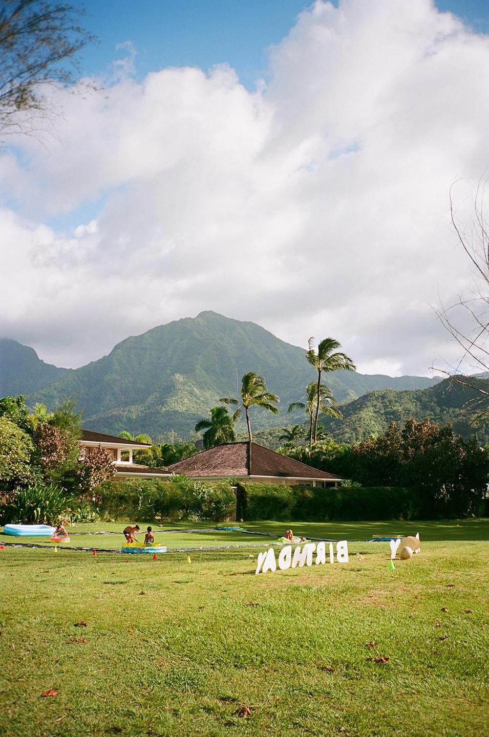 A large green lawn with children playing and a "BIRTHDAY" sign in the foreground, with houses, palm trees, and mountains in the background.