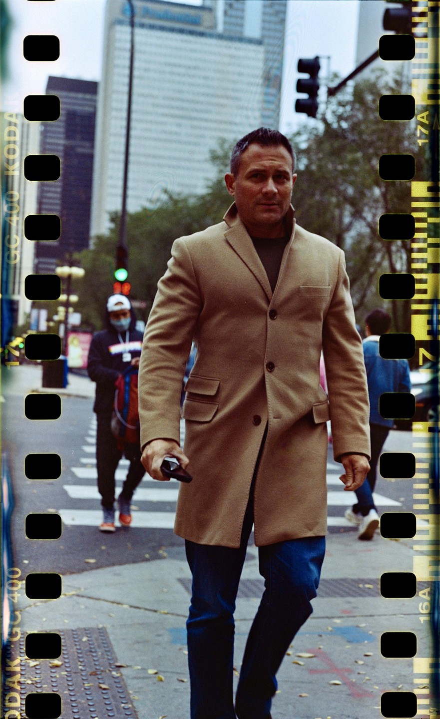 A man in a tan coat walks across a city street with tall buildings in the background.