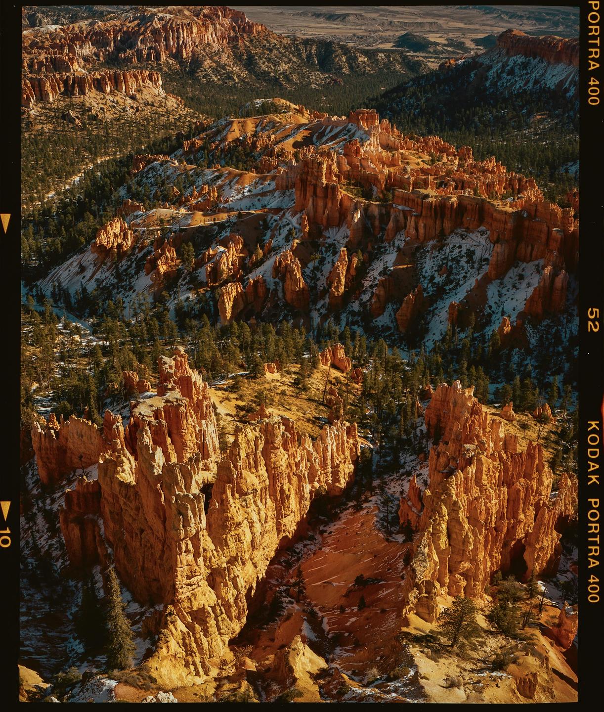 An aerial view shows a vast canyon landscape with orange rock formations and scattered pine trees, with patches of snow on the ground.