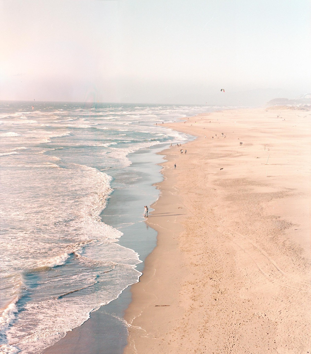 A wide shot shows a beach with waves crashing on the shore and people walking on the sand.
