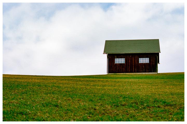 A small wooden house with a green roof sits on a grassy hill under a cloudy sky.