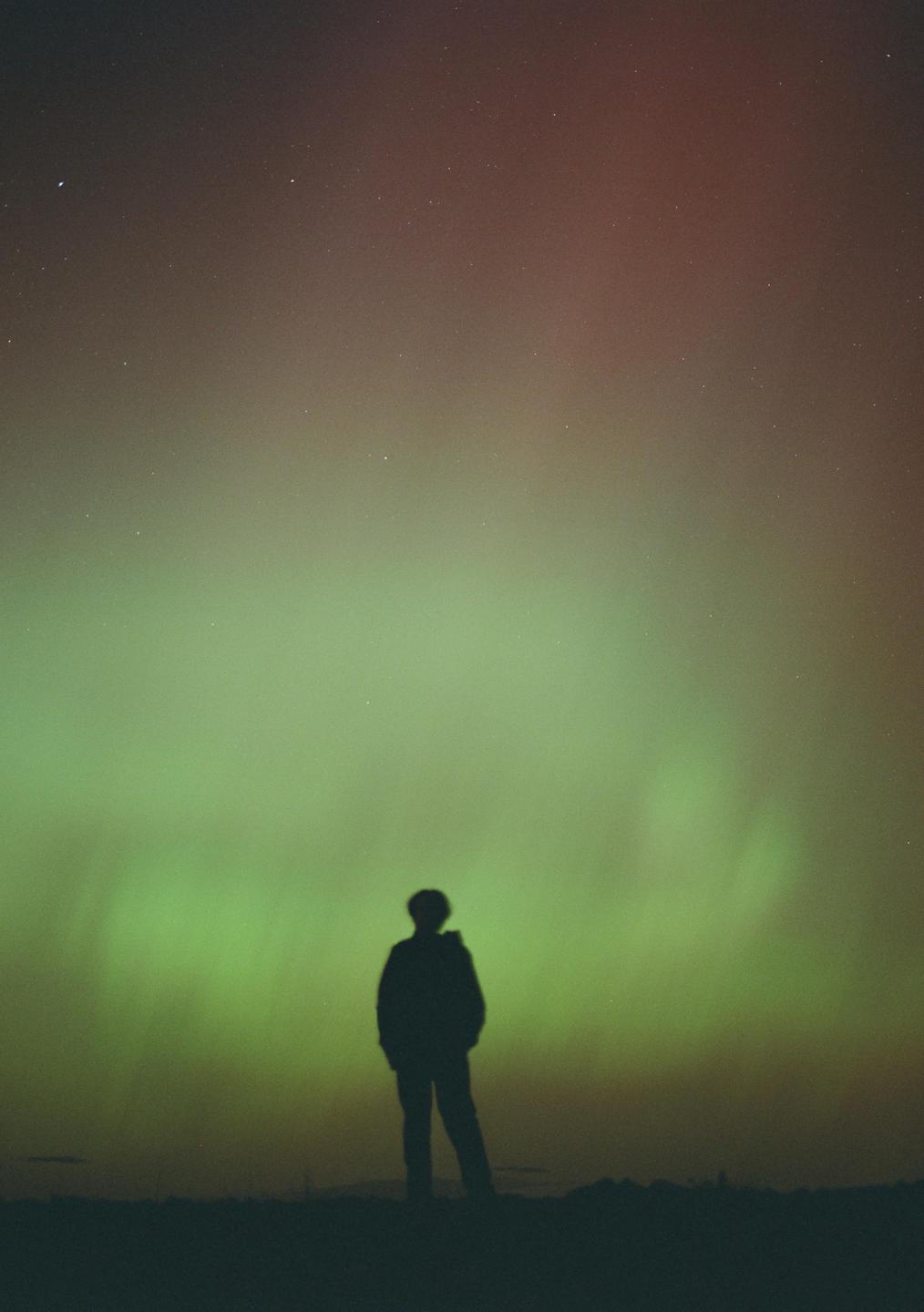 A person stands silhouetted against a night sky filled with the aurora borealis.