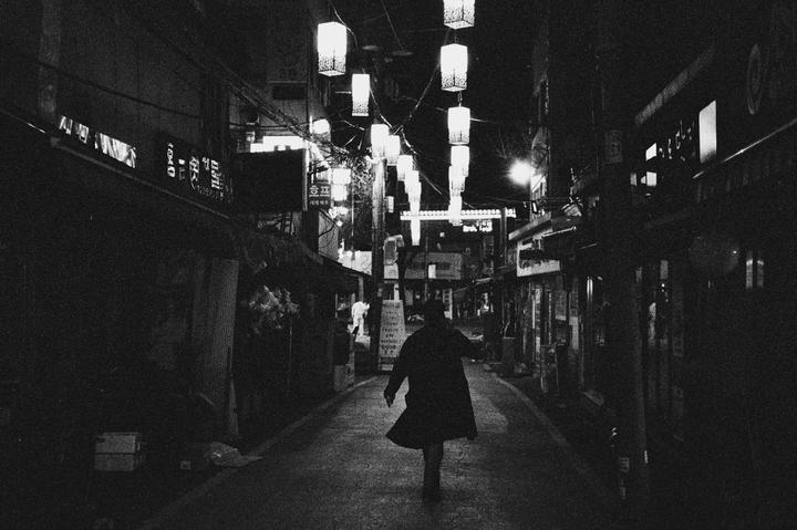 A person walks down a narrow street at night, illuminated by hanging lanterns and storefront lights.