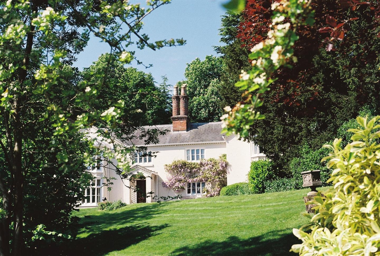 A white house with a wisteria vine growing on its facade is nestled among green trees and a grassy lawn.