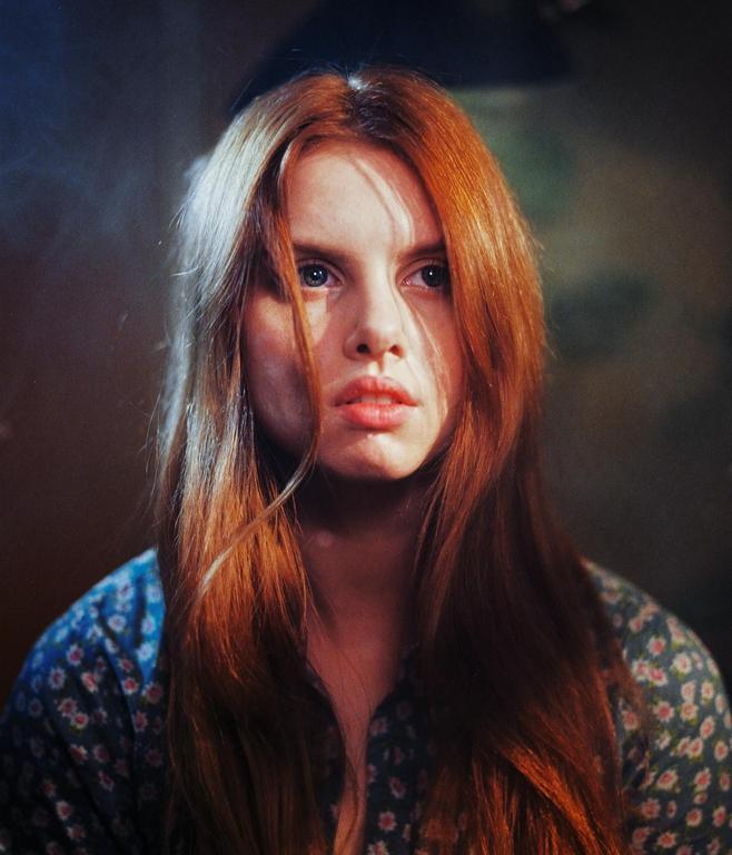 A woman with long red hair and a floral shirt looks directly at the viewer.