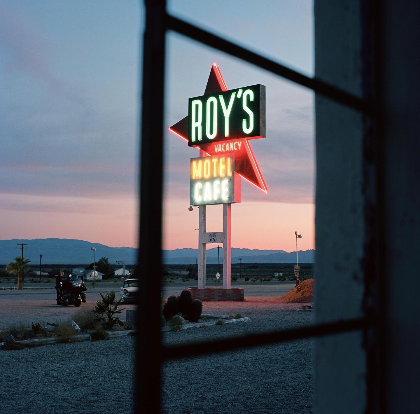 A neon sign for Roy's Motel and Cafe with a vacancy sign is visible at dusk, framed by a window.