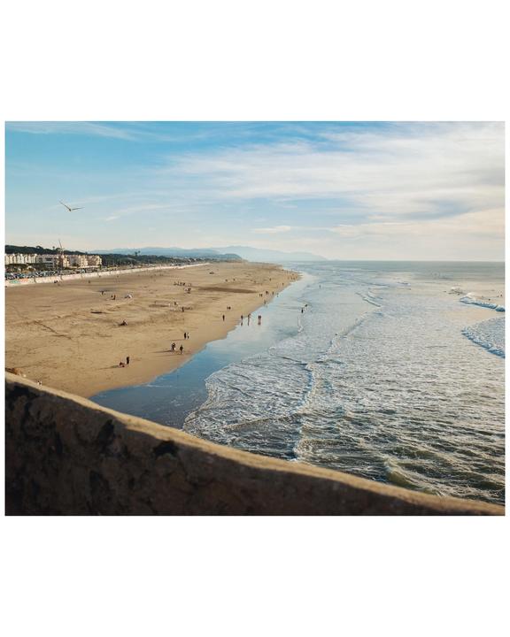 A wide view of a beach with people walking along the shore and waves crashing, with buildings and mountains in the distance.