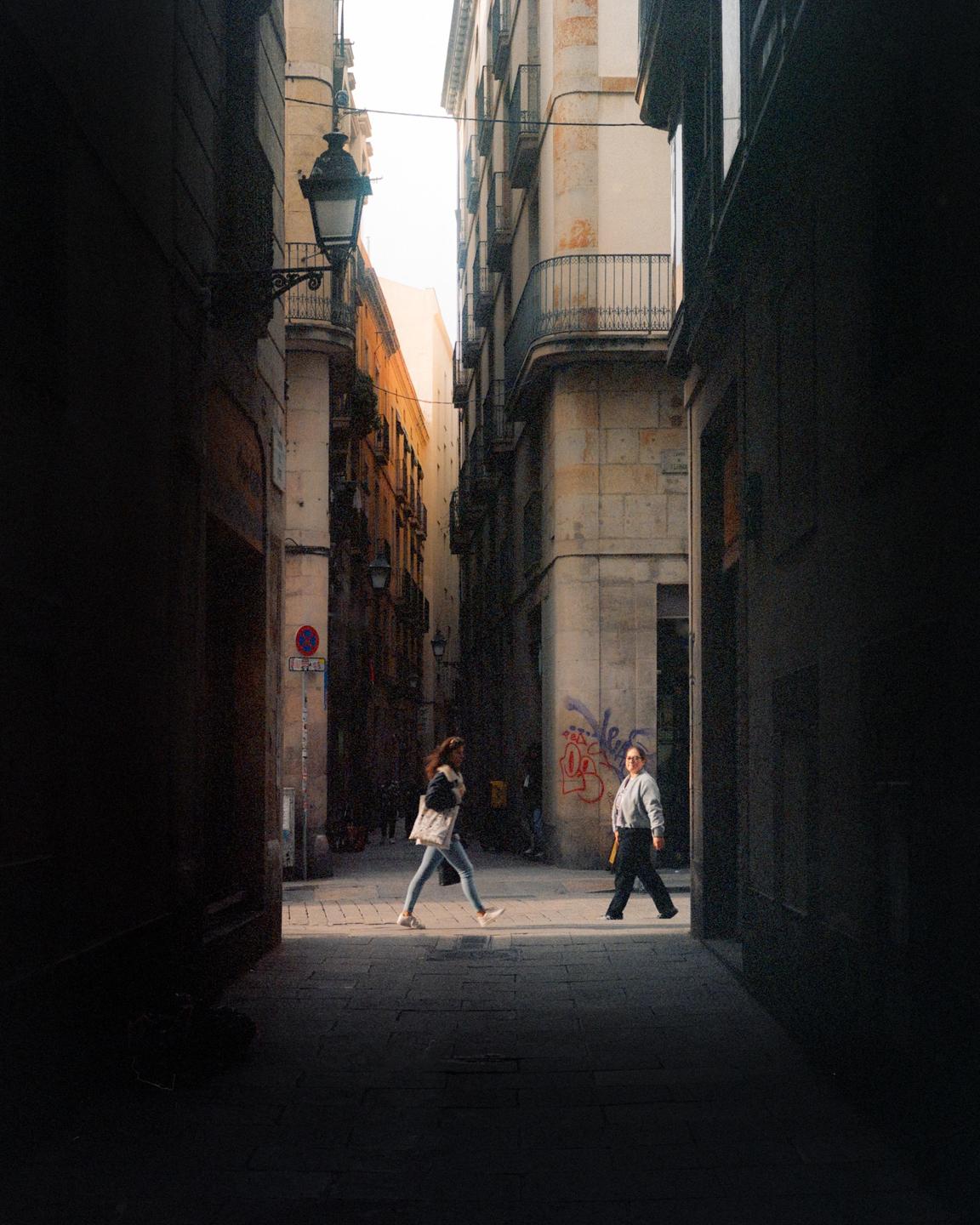 Two women walk through a narrow street with tall buildings on either side, illuminated by a patch of sunlight.