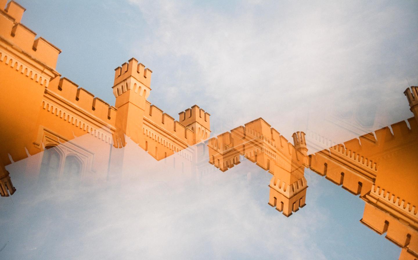 A double exposure shows the orange facade of a building with battlements against a blue sky.