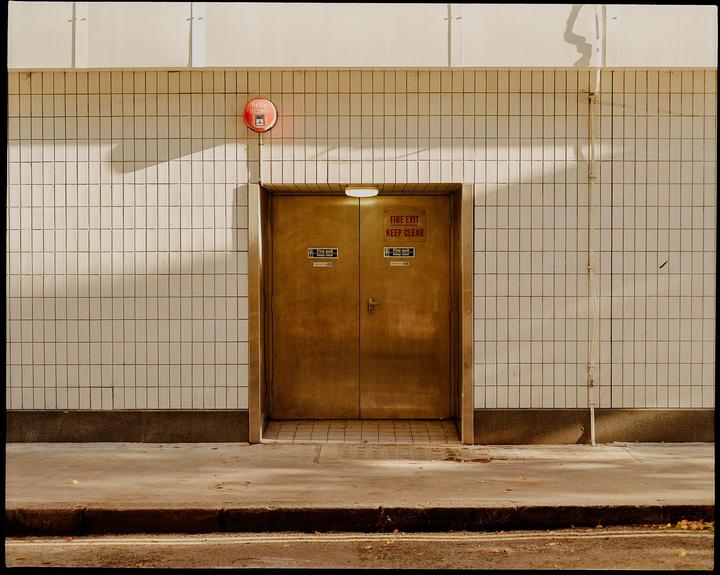 A fire exit door is set into a tiled wall with a security alarm above it, casting shadows on the wall.