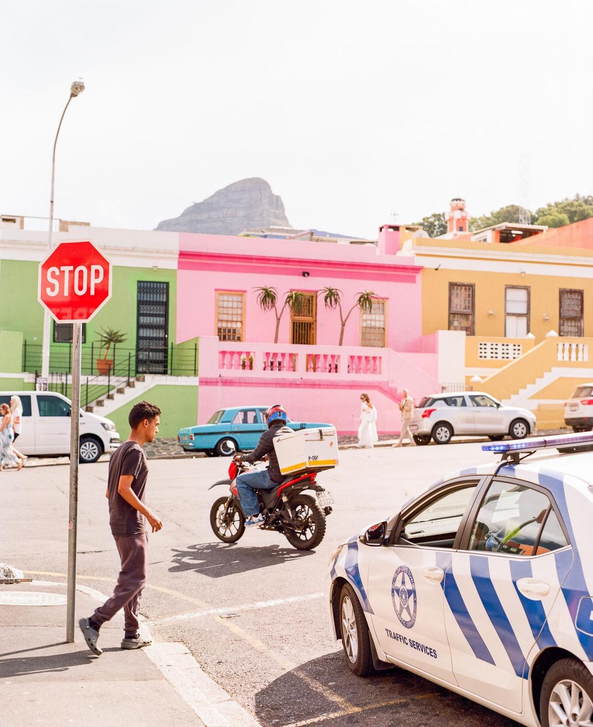 A boy walks past a stop sign and a traffic services car with colorful houses and a mountain in the background.