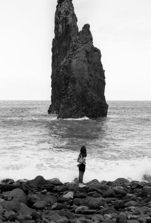 A girl stands on a rocky beach looking out at a large rock formation in the ocean.