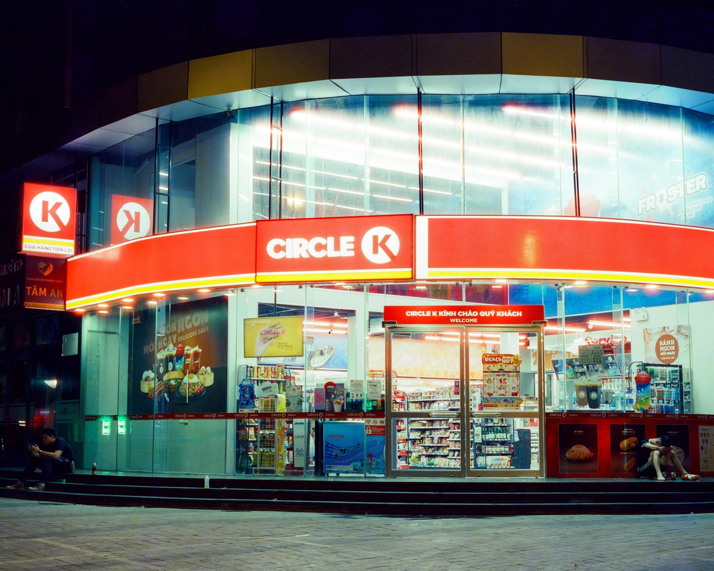 A Circle K convenience store is brightly lit at night with two men sitting outside.