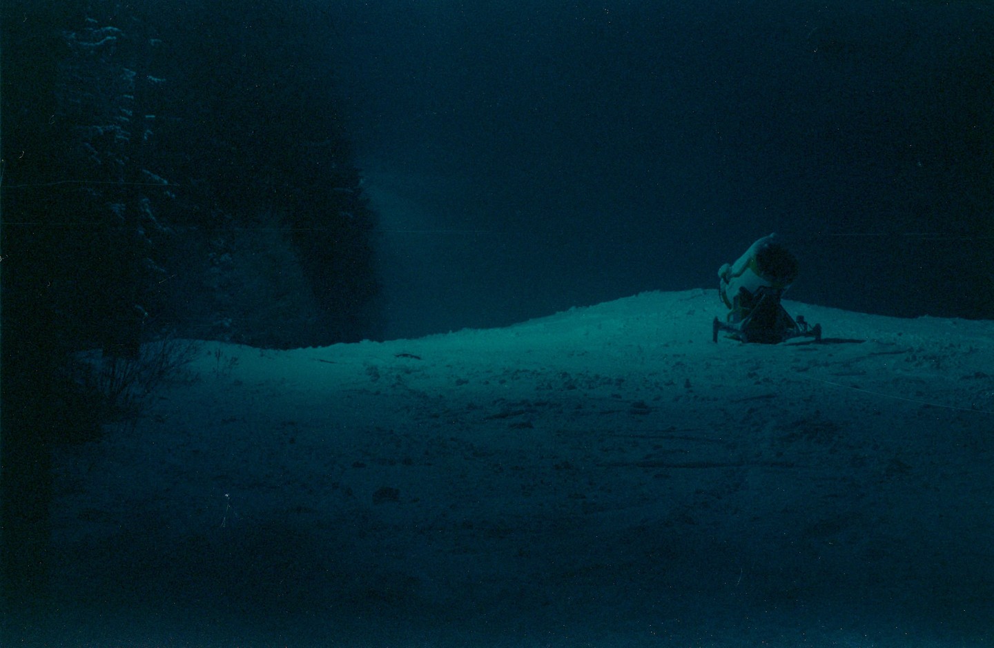 A snow cannon sits on a snow-covered hill at night, with trees visible in the dark background.
