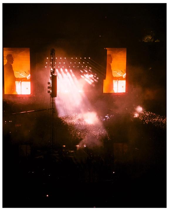 A large crowd of people are visible at a night concert with bright stage lights and two large screens showing a man.