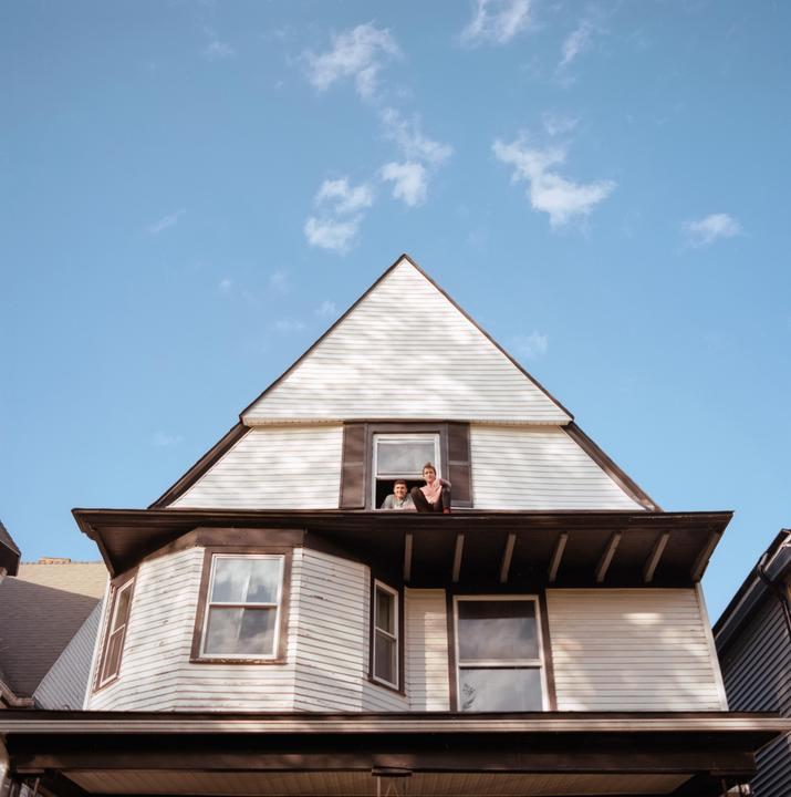 A couple is sitting in an open window on the top floor of a white house with a blue sky and clouds above.