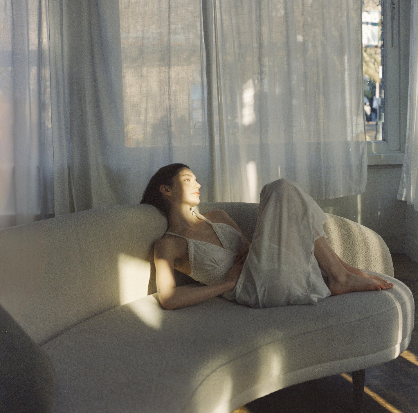 A woman in a white dress is lying on a sofa in a sunlit room.