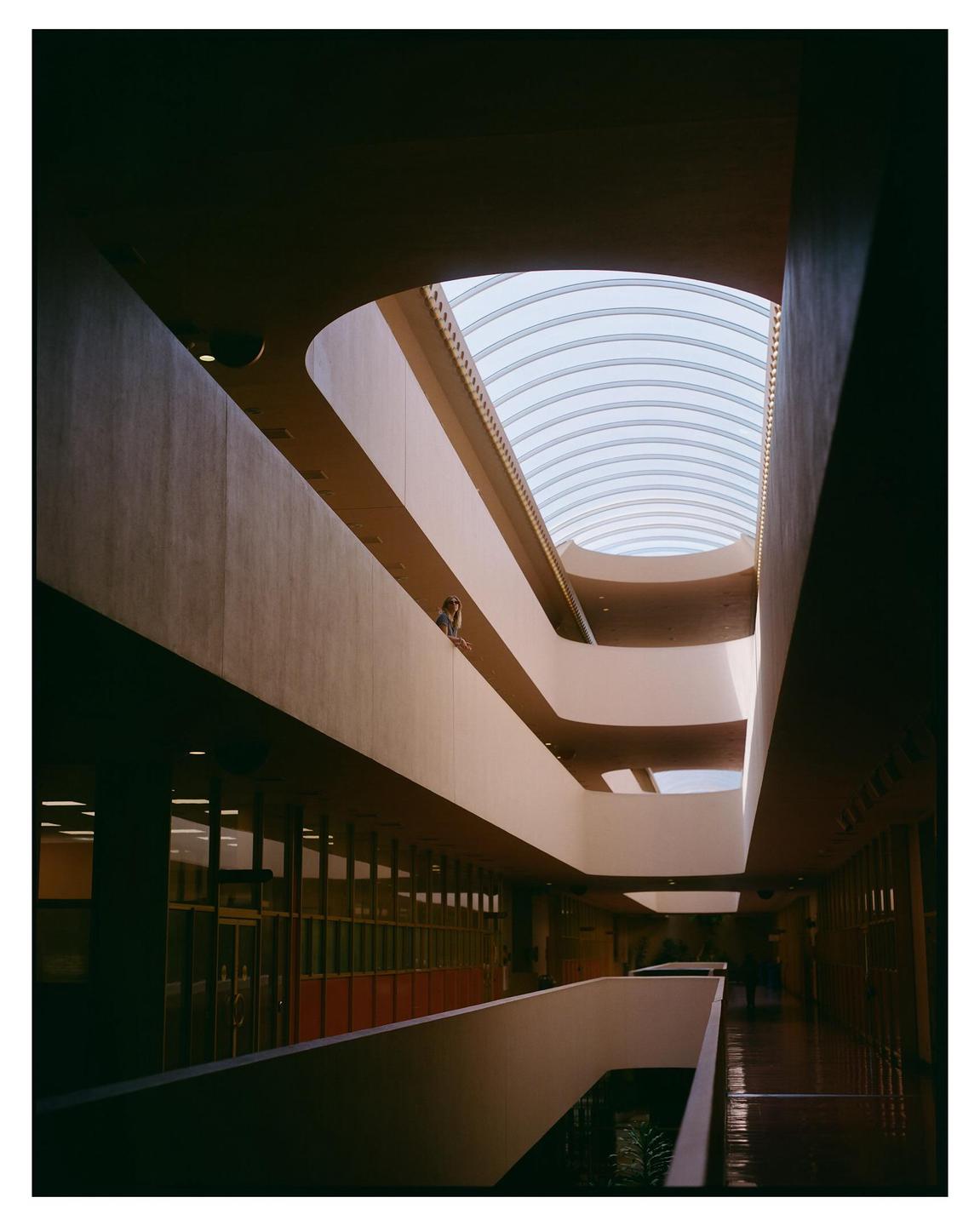A woman stands on a balcony overlooking multiple levels of an indoor structure with a large skylight.
