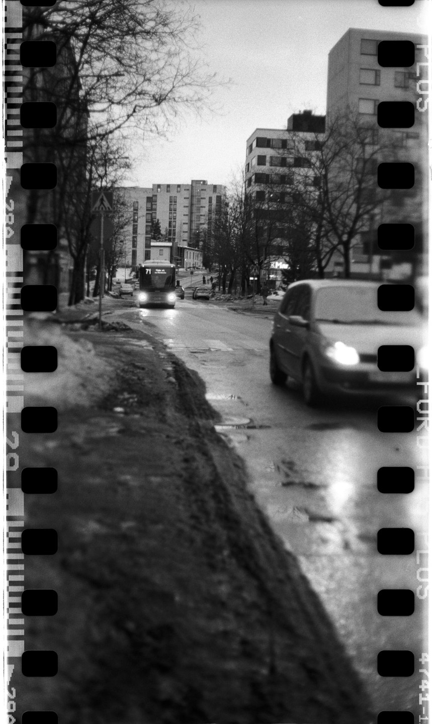 A bus and a car drive on a wet street lined with bare trees and buildings.
