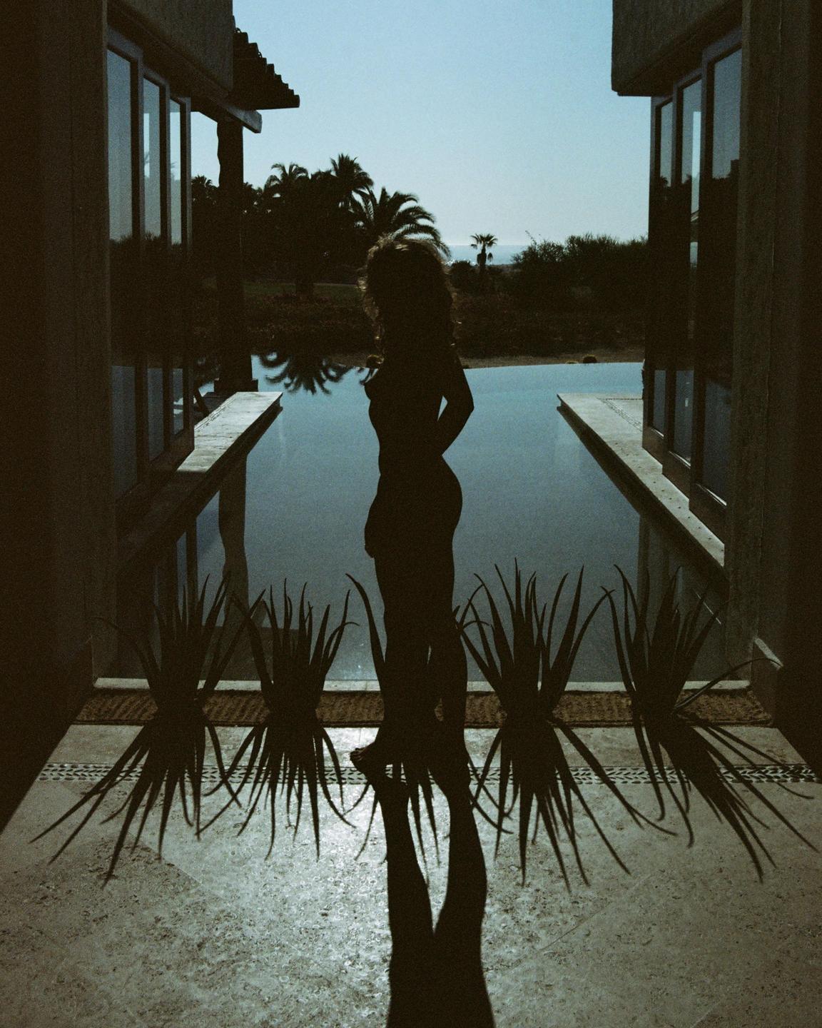A woman stands silhouetted against a pool with palm trees and the ocean in the background.