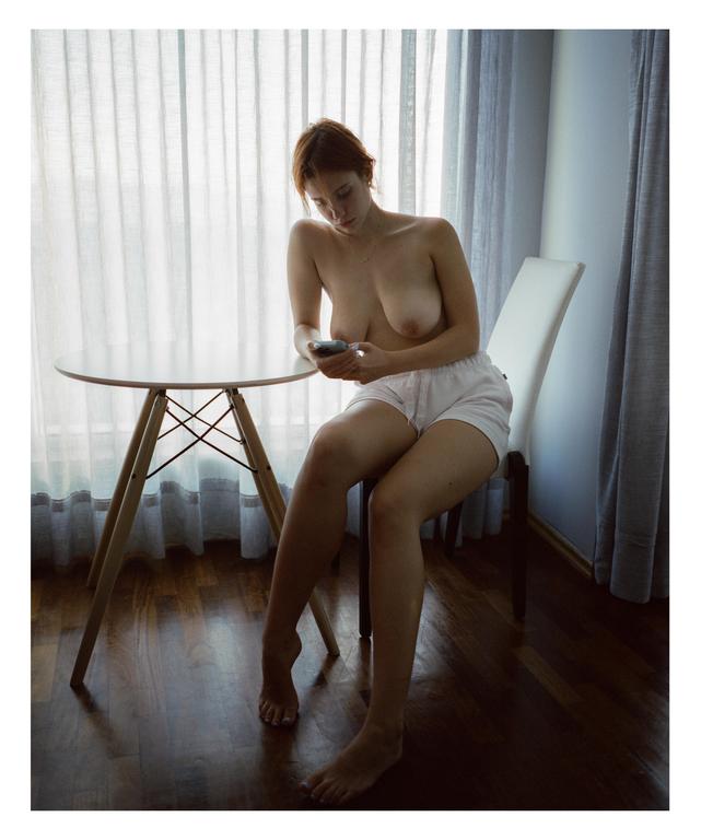A woman sits topless at a table, looking at her phone.