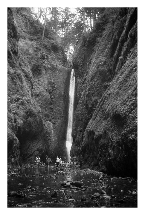 A group of people stand in a rocky stream at the base of a tall waterfall cascading between mossy cliffs.