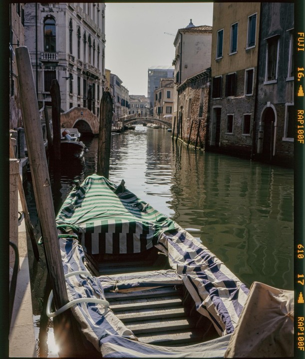 A gondola covered with striped fabric is moored in a canal lined with old buildings and bridges.