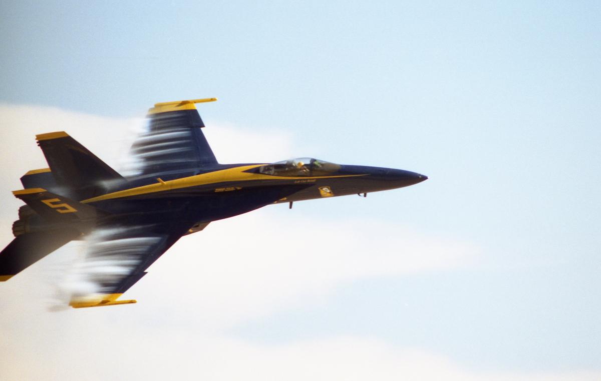 A blue and yellow jet flies through the sky, leaving a trail of vapor behind its wings.