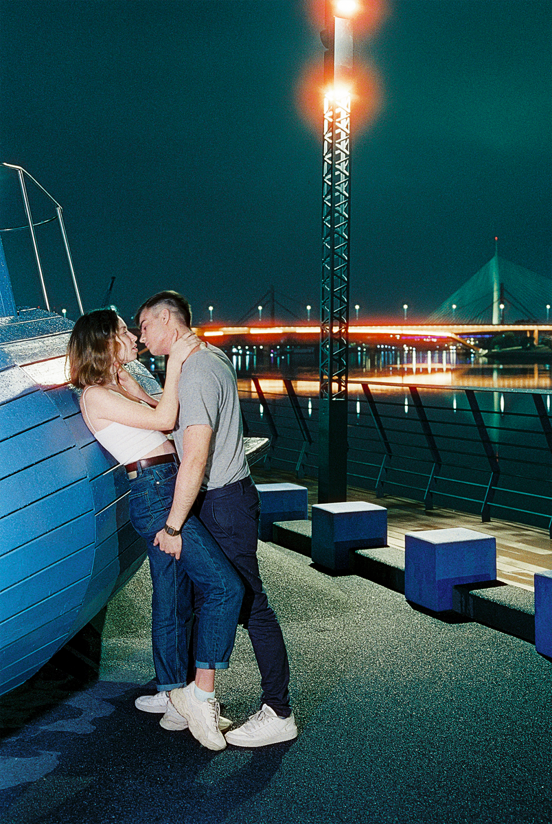 A couple embraces next to a blue structure on a riverside promenade at night, with a bridge and city lights in the background.