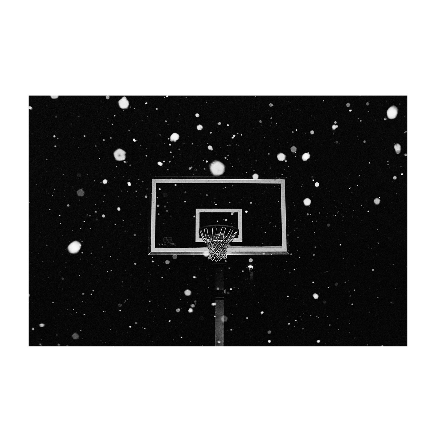 A basketball hoop and backboard are visible at night with snow falling.