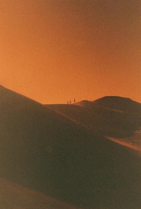 Three people stand silhouetted on a sand dune against an orange sky.