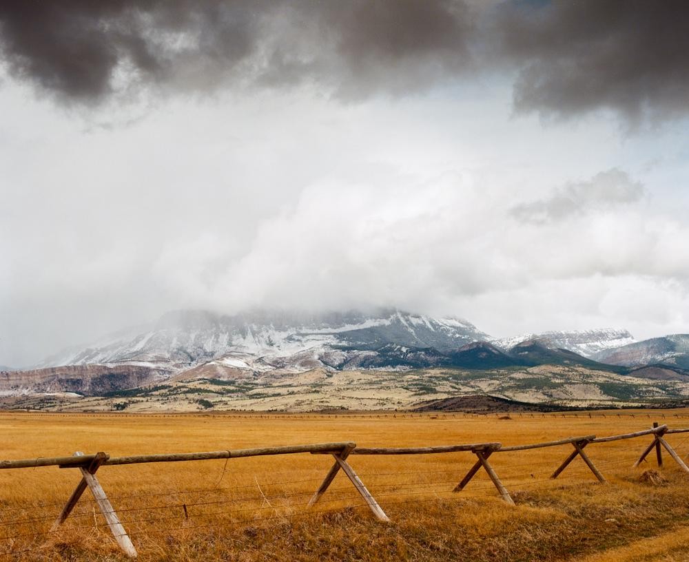 A wooden fence stretches across a golden field with snow-capped mountains in the background under a cloudy sky.