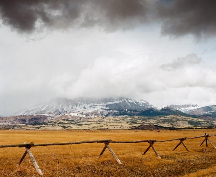 A wooden fence stretches across a golden field with snow-capped mountains in the background under a cloudy sky.