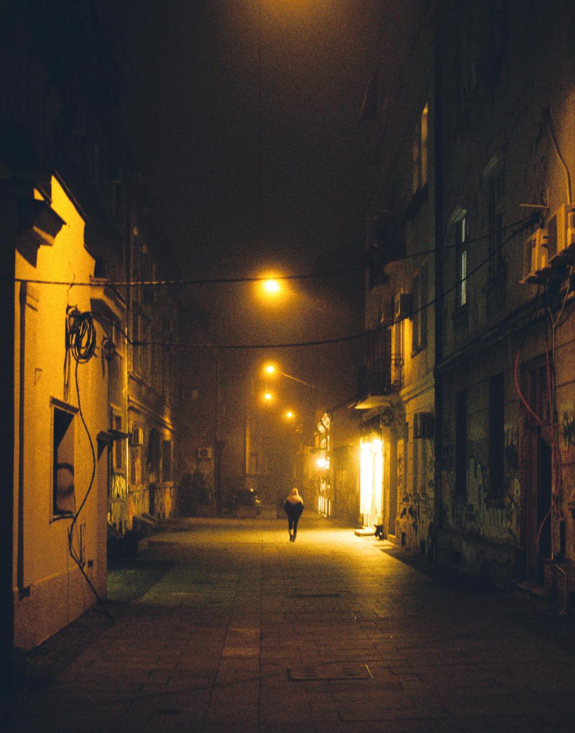 A person walks down a narrow street at night, illuminated by streetlights and building lights.