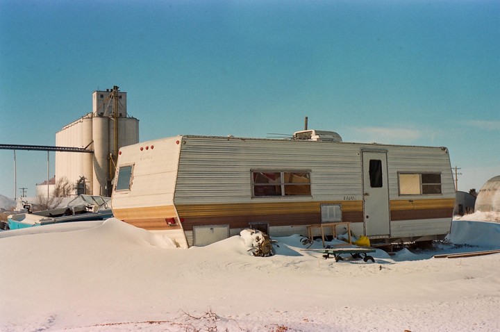 A white and brown camper is parked in a snow-covered field with grain silos in the background.