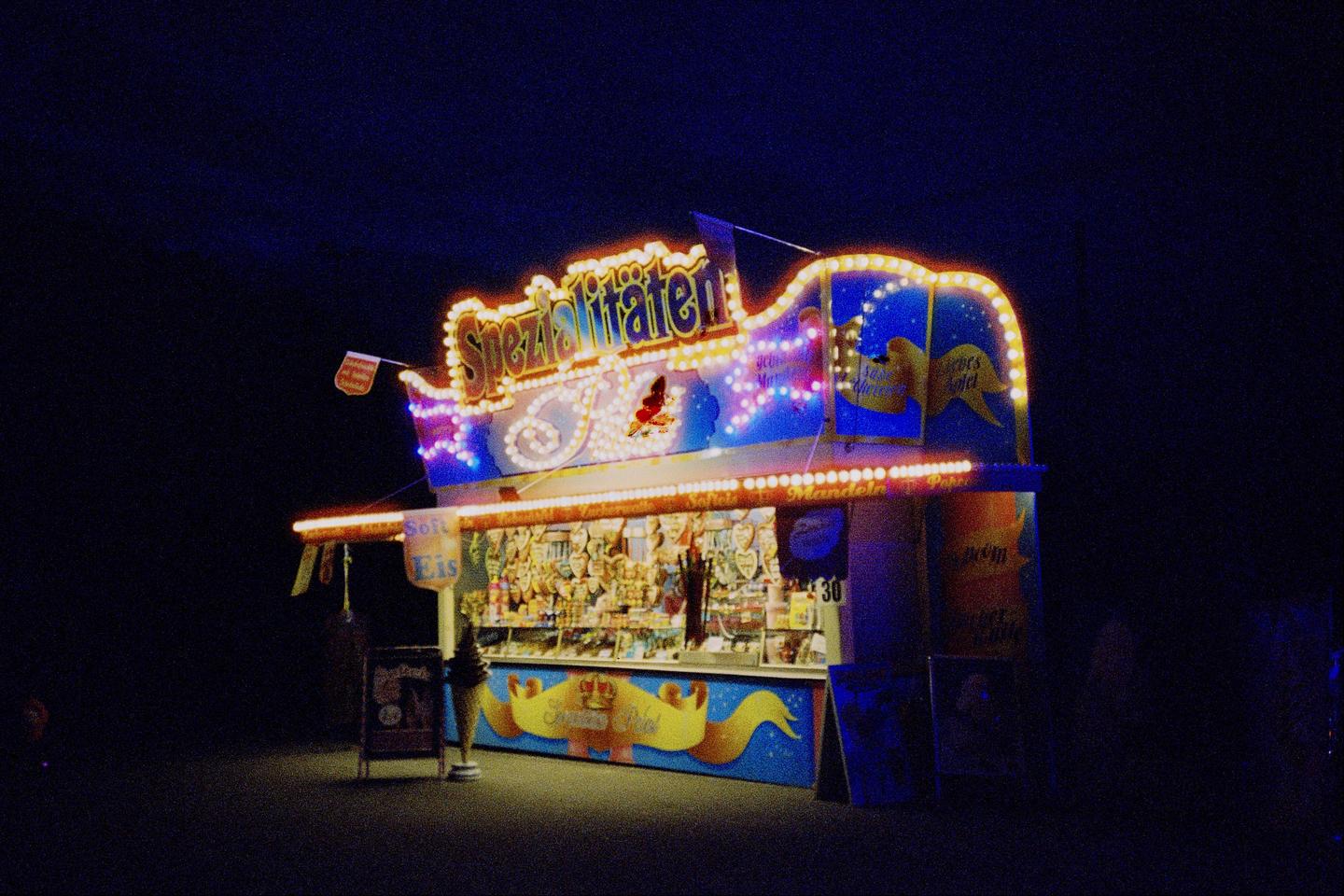A brightly lit food stall with a sign that reads "Spezialitäten" stands out against a dark night sky.