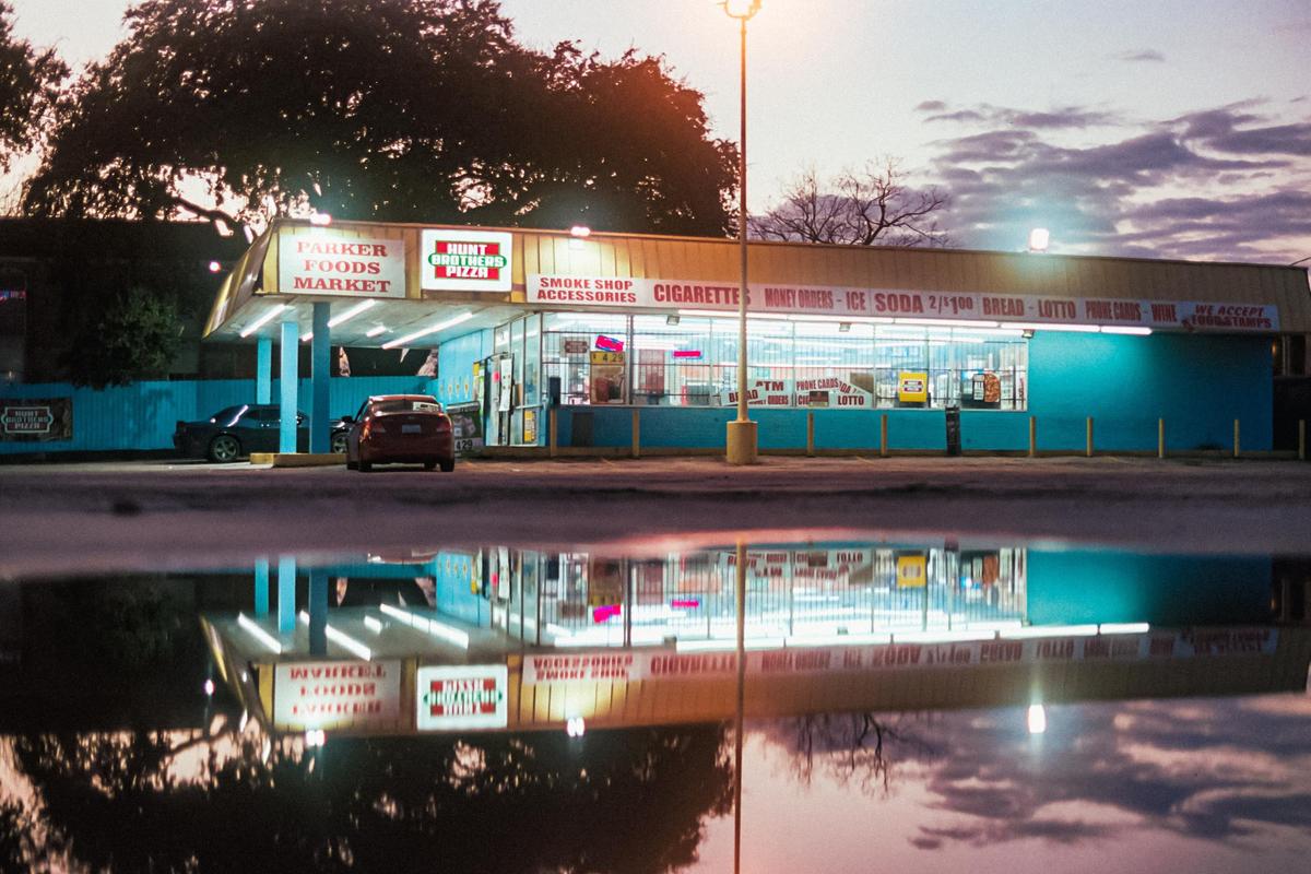 A convenience store with a bright blue facade and illuminated signs is reflected in a puddle at dusk.
